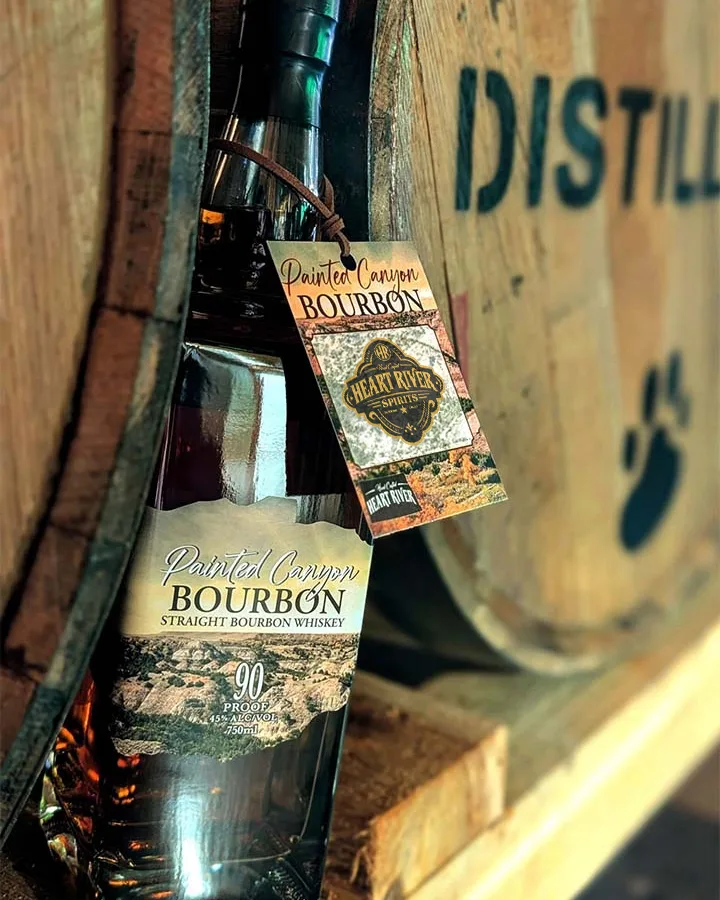 Painted Canyon Bourbon bottle with a Heart River Spirits hang tag, resting against a Big Dog Distillery barrel