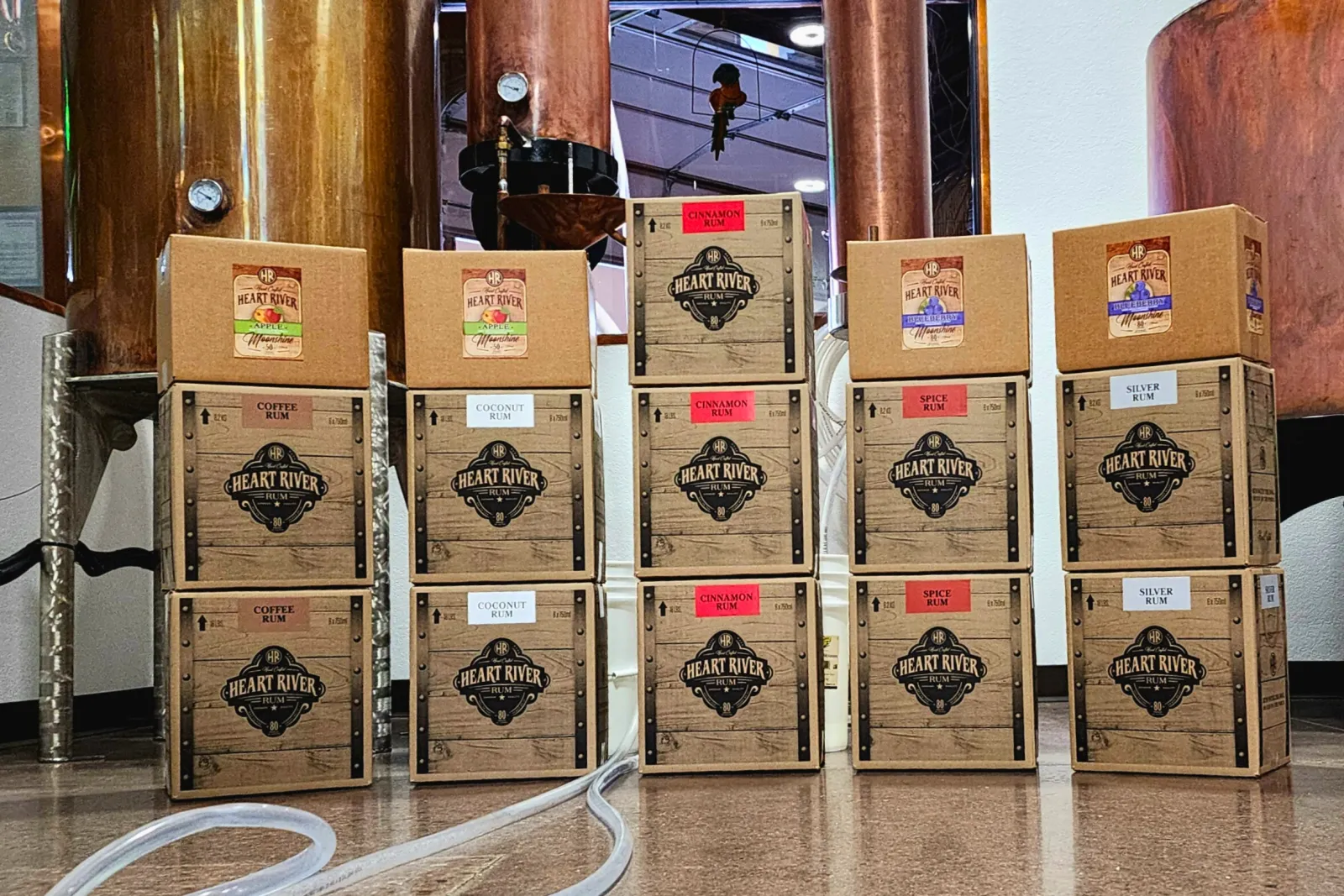 Cases of Heart River Spirits stacked in front of the copper stills at Big Dog Distillery